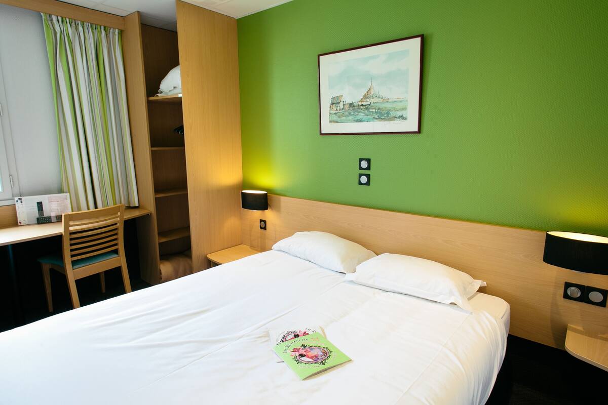 A hotel room with a bed, a desk, and a chair. There is a painting on the wall and a lamp on the nightstand.