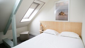 Ein Hotelzimmer mit schrägen Wänden, einem Bett mit weißer Bettwäsche, einem kleinen Nachttisch mit Lampe und einem gerahmten Bild an der Wand.