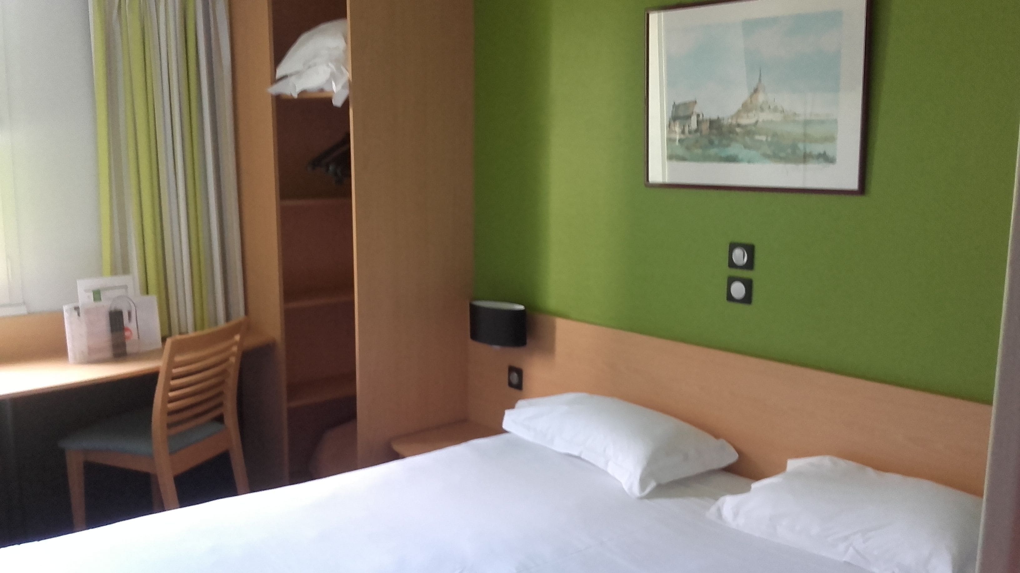 A hotel room with a bed, a desk, a chair, a wardrobe, and a painting on the wall.