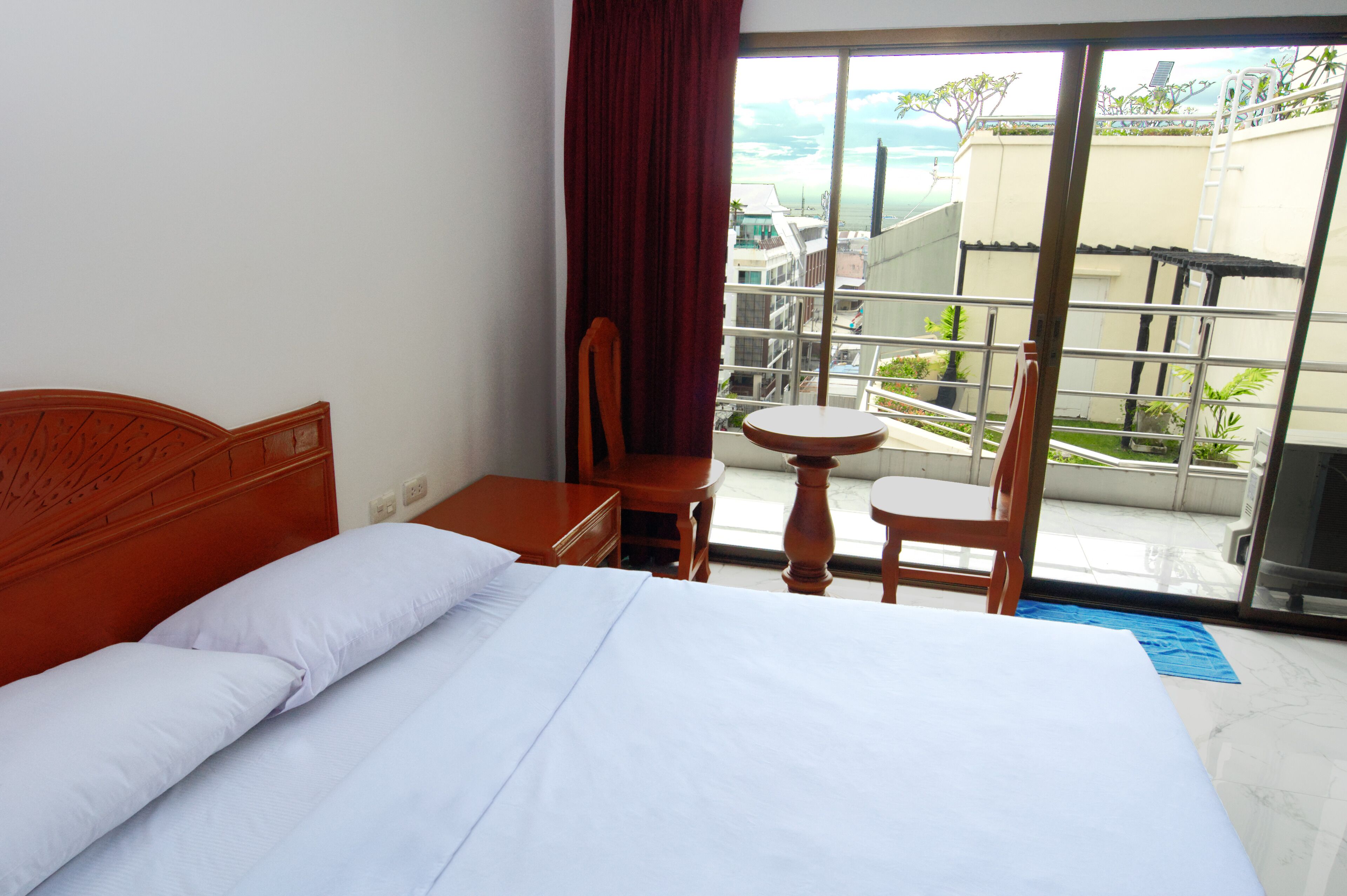 A hotel room with a bed, a wooden nightstand, a small round table, two chairs, and a large sliding glass door leading to a balcony with a view of buildings and greenery.