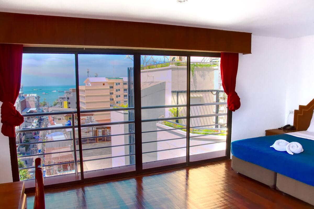 A hotel room with a large sliding glass door leading to a balcony, a bed with a blue bedspread and two towels, and a wooden floor.