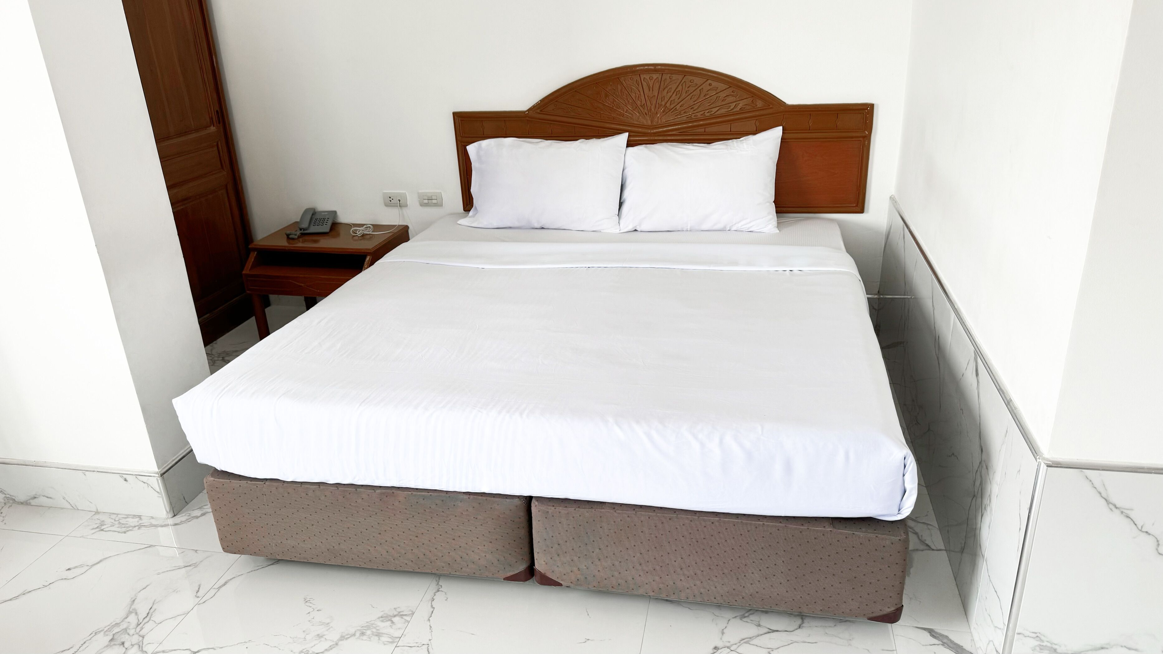 A neatly made bed with a wooden headboard, a bedside table with a phone, and a wall socket.