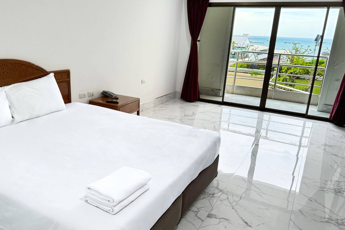 A hotel room with a large bed, a wooden headboard, a nightstand, and a view of the sea through a sliding glass door.