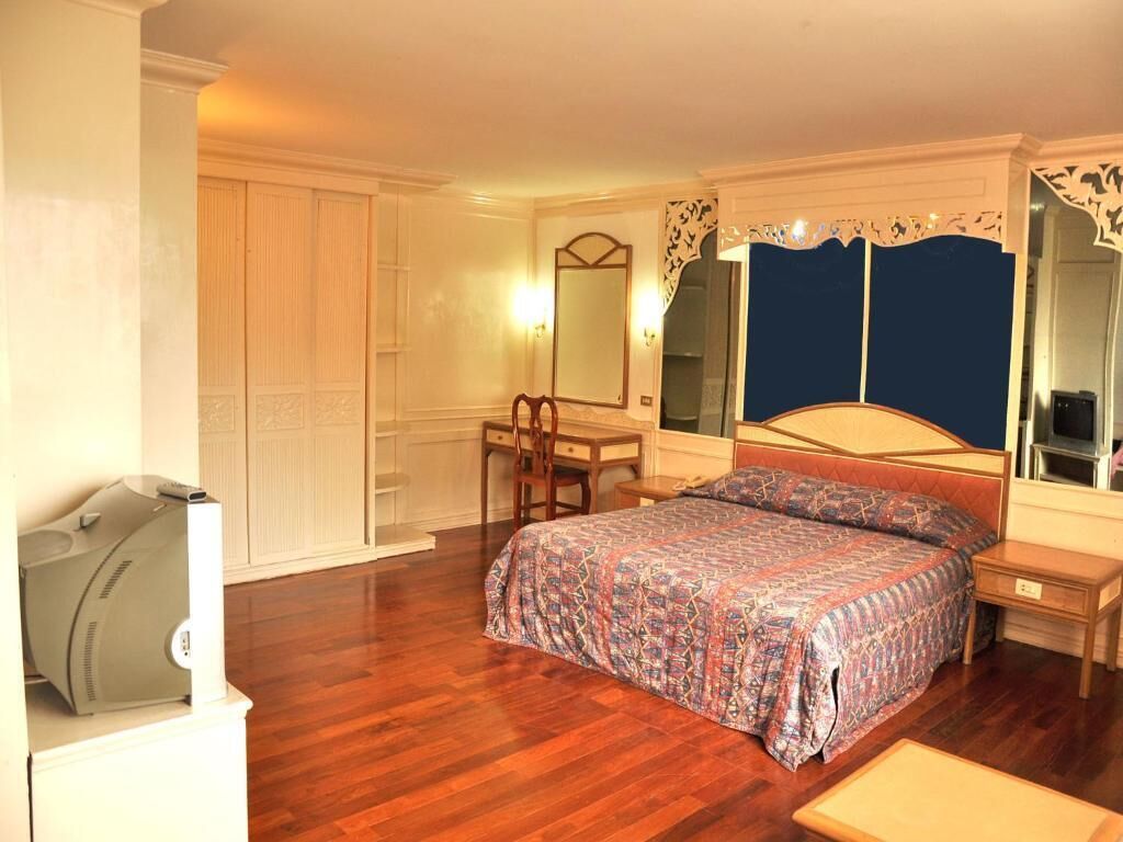 A bedroom with a wooden floor, a bed with a patterned bedspread, a bedside table, a chair, a desk, a mirror, and a wardrobe.