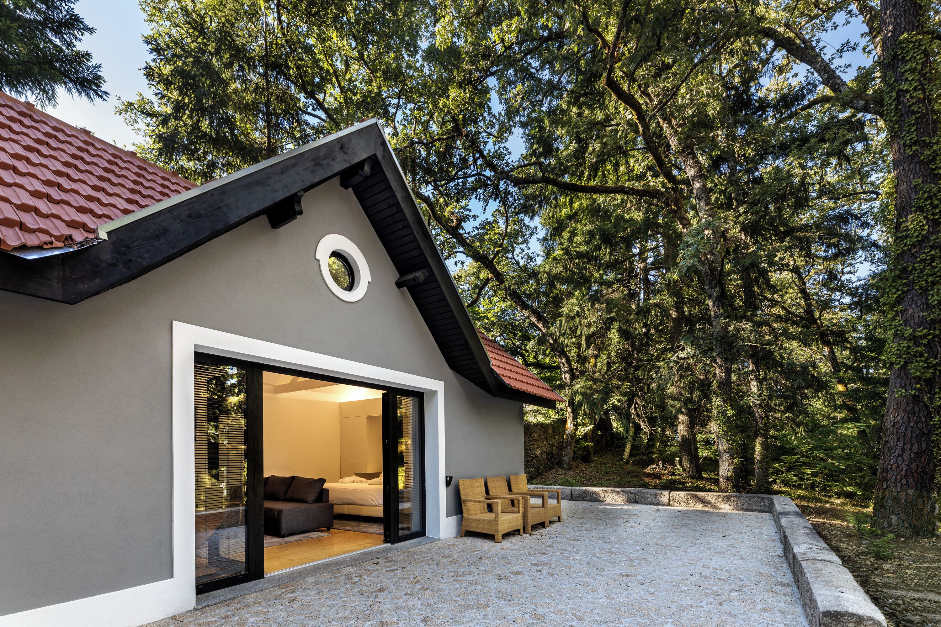 Una casa moderna con techo inclinado, ventana circular y puerta de vidrio que da a un dormitorio con cama y sillón.