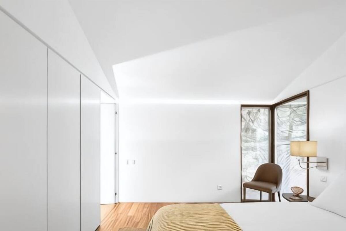A modern bedroom with a bed, a chair, a lamp, and a view through a window.
