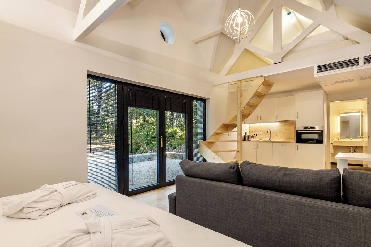 A modern hotel room with a large bed, a sofa, a staircase, and a kitchen area.