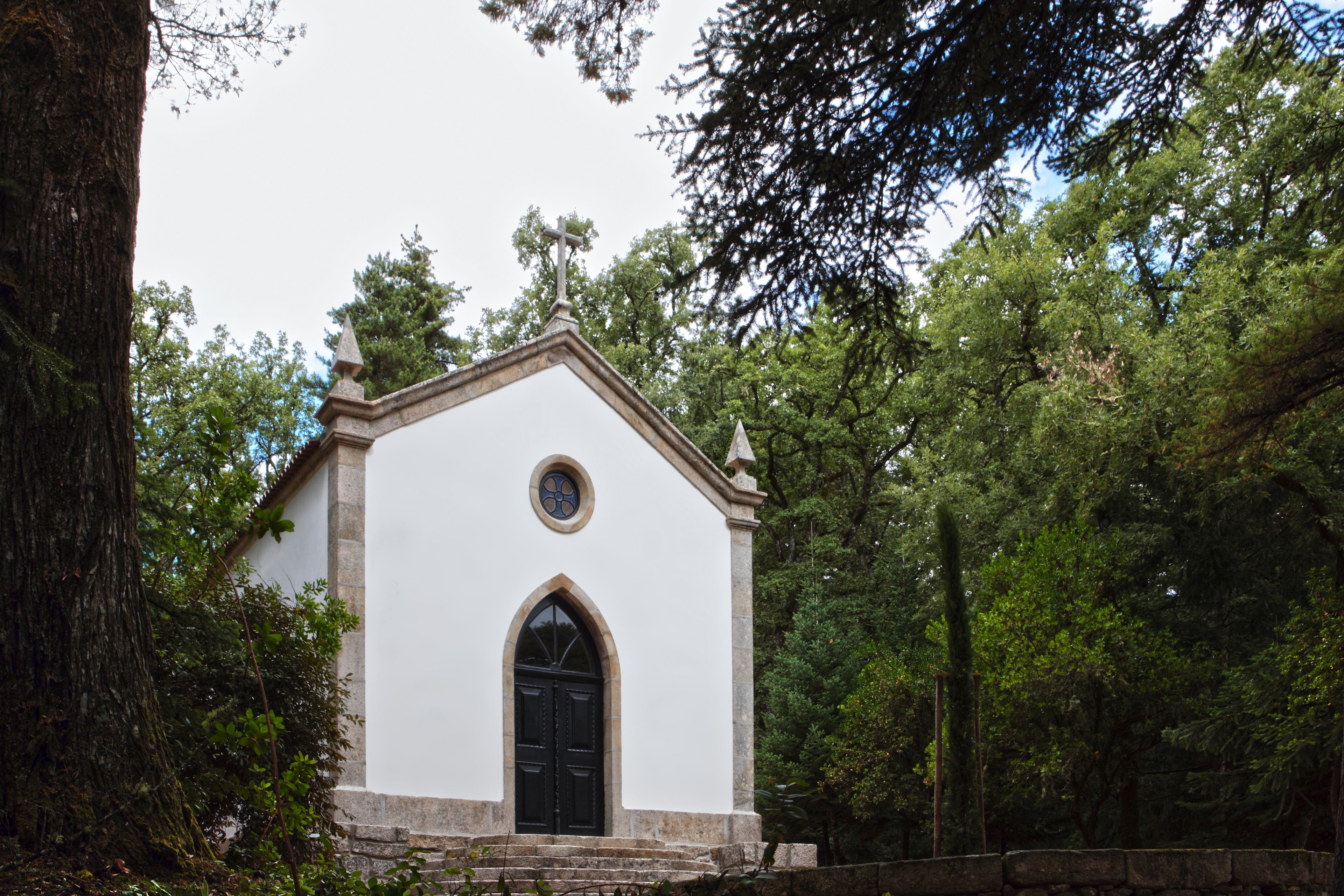 A small white church with a black door and a circular window, surrounded by trees.