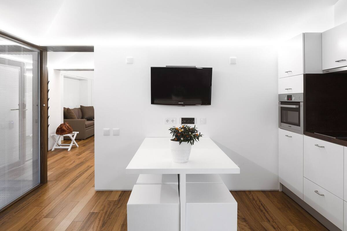 A modern kitchen with white cabinetry, a built-in oven, and a flat-screen TV mounted on the wall.