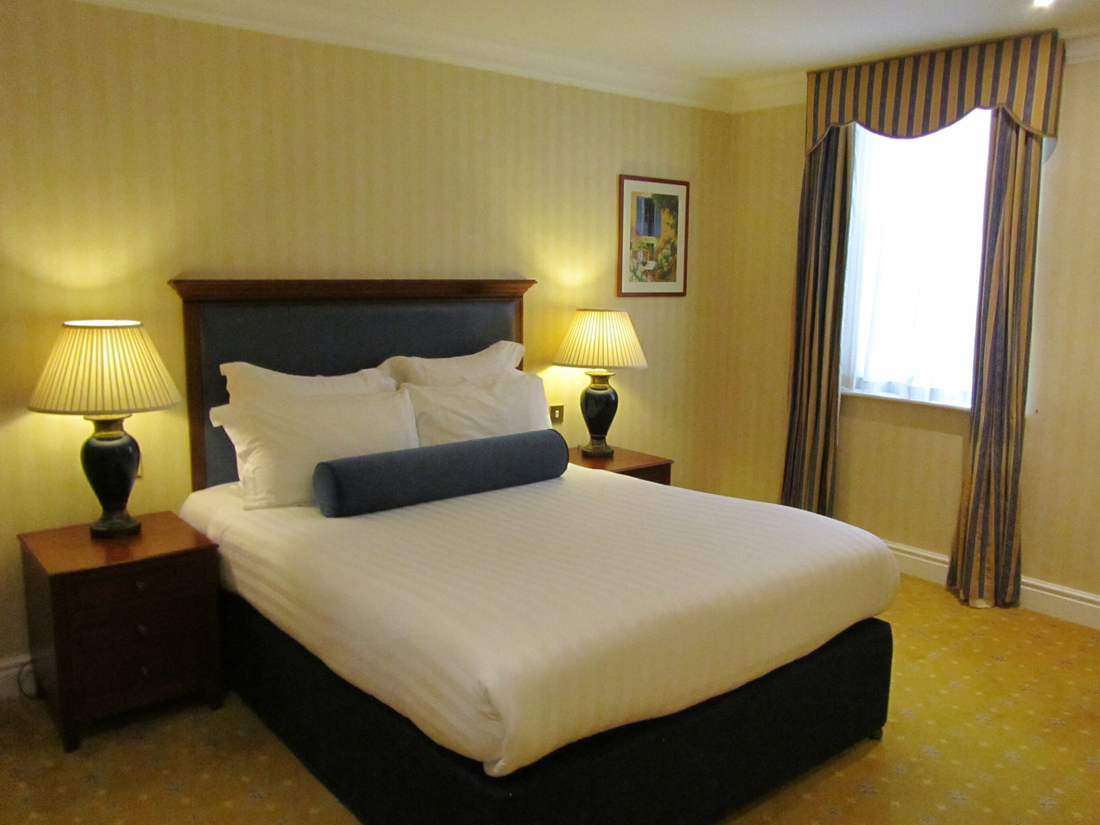 A neatly made bed with white linens and a blue accent pillow, flanked by two bedside tables with lamps, in a room with a large window and curtains.