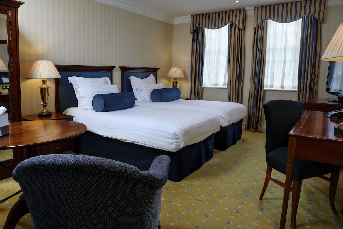A hotel room with a double bed, two bedside tables with lamps, a desk with a chair, and a large mirror.
