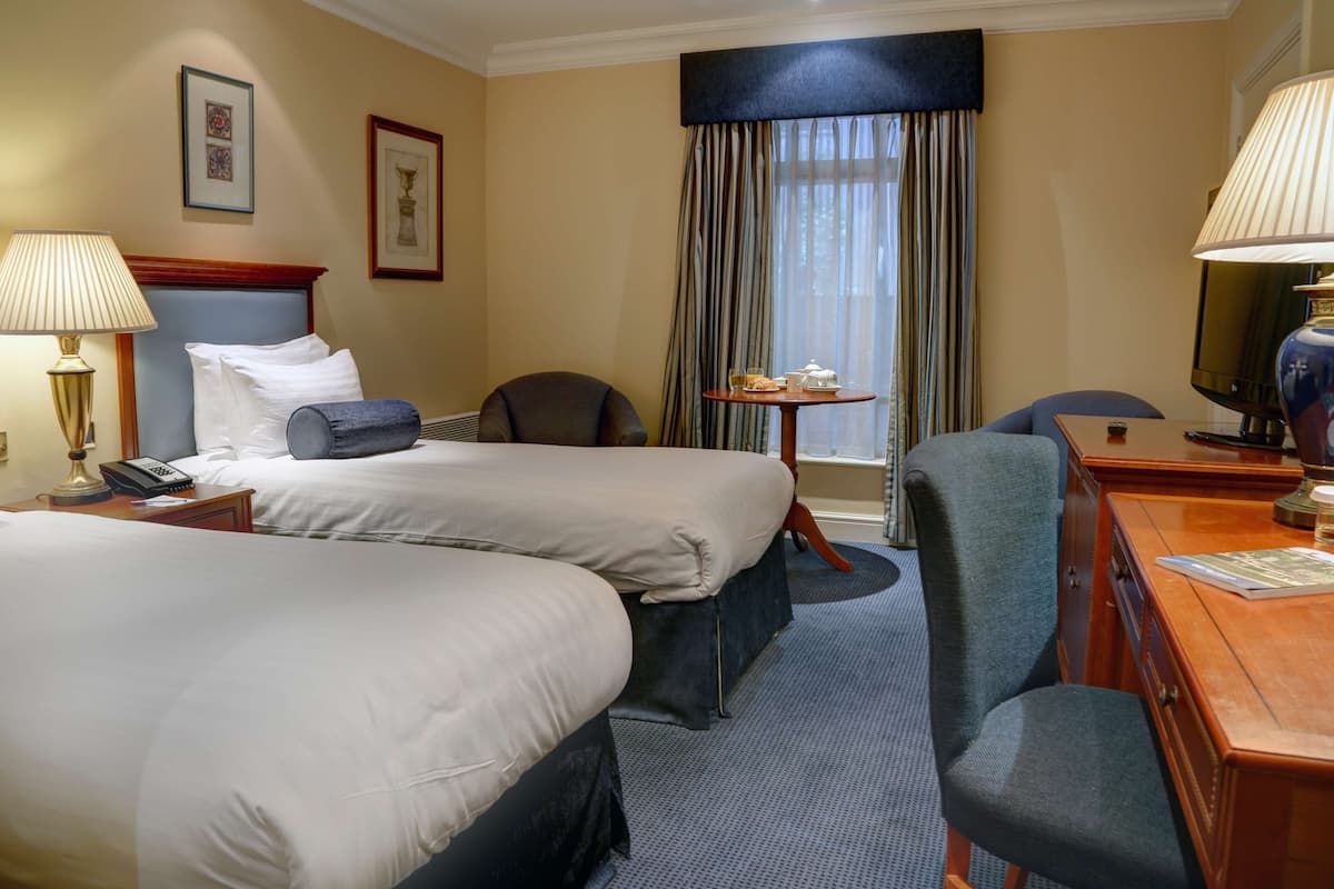 A hotel room with two beds, a desk, a chair, a lamp, a television, and a window with curtains.