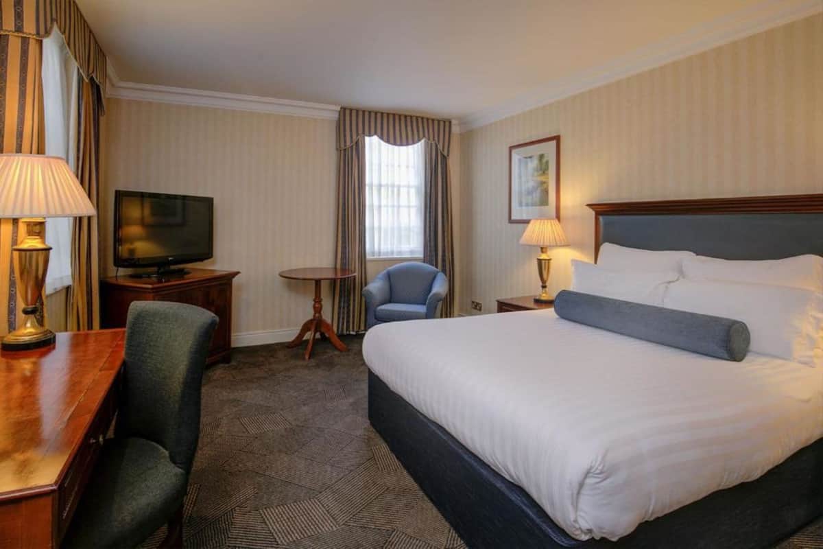 A hotel room with a large bed, a desk with a lamp, a television, a chair, and a small table.