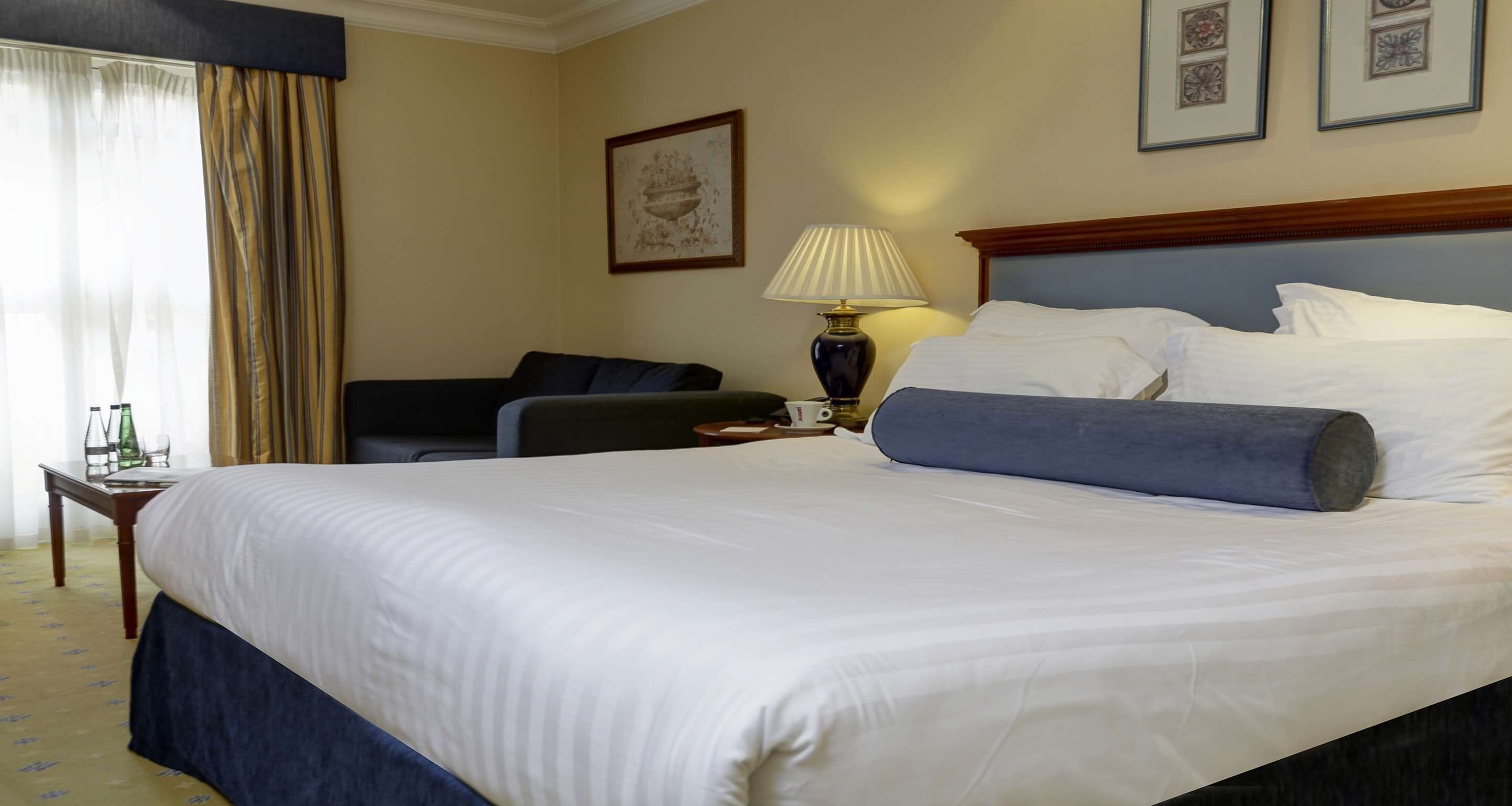 Una camera d'albergo con un letto grande, un comodino con una lampada, una sedia e un quadro appeso al muro.