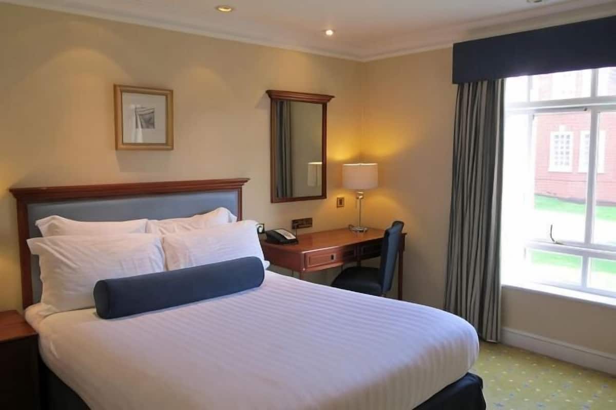A hotel room with a bed, a desk, a chair, a lamp, a mirror, and a window with a view of a building.