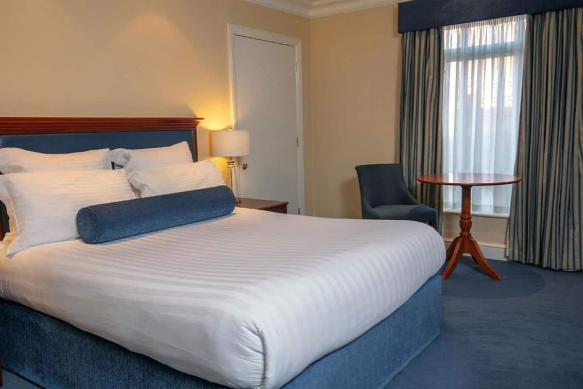 A hotel room with a large bed, a chair, a small table, and a window with curtains.