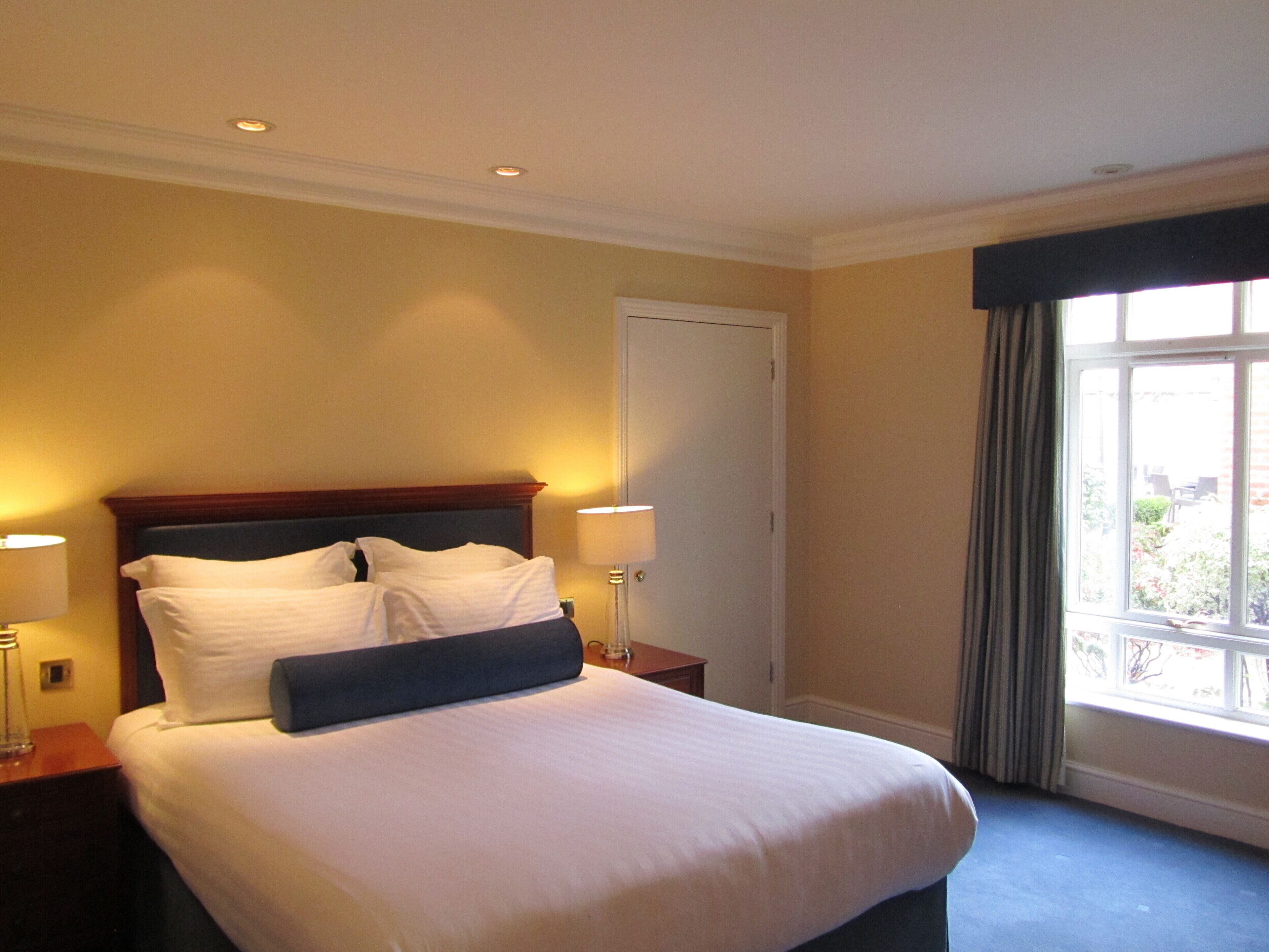 A hotel room with a large bed, two bedside lamps, a window with curtains, and a door.
