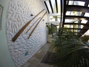A room with a textured white wall, wooden bat hanging, a potted plant, and a window with a view of palm trees. - Pousada do Capitão (Arraial do Cabo)