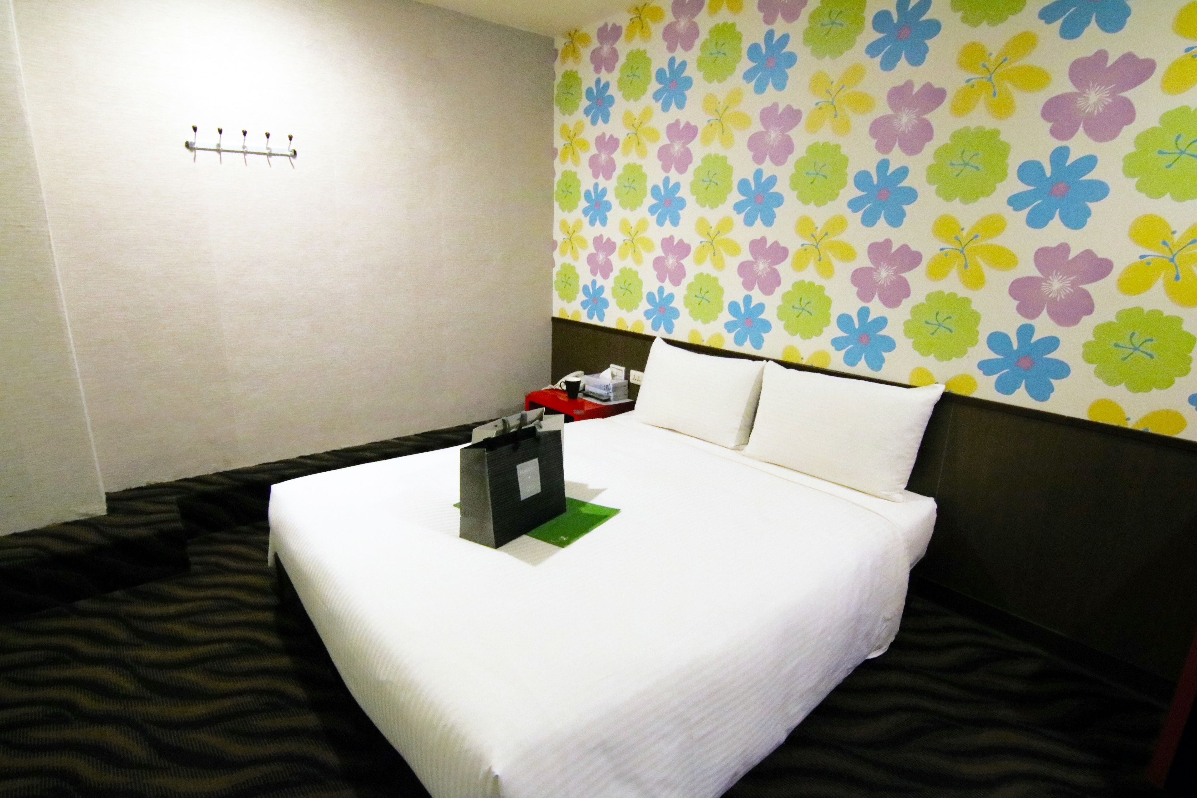 A hotel room with a bed, a headboard, a wall with a floral pattern, and a small table with a bag and a red object.