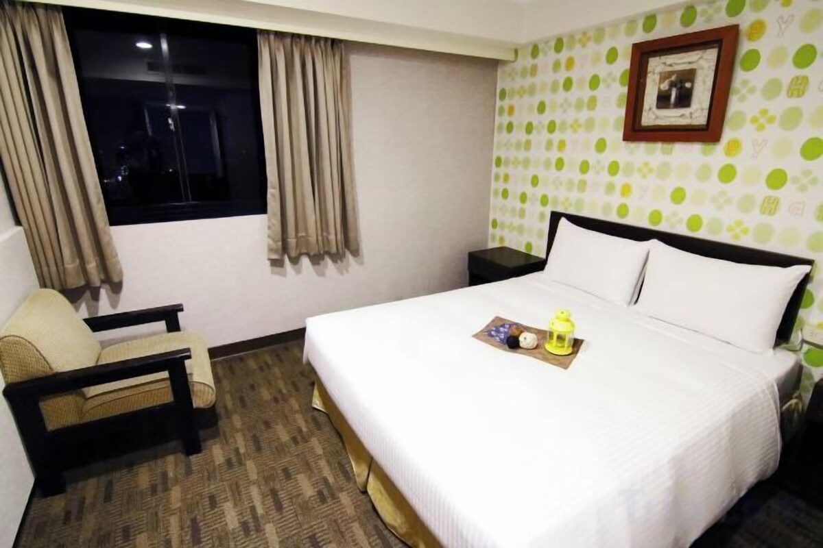 A hotel room with a bed, a chair, a nightstand, and a window with curtains.