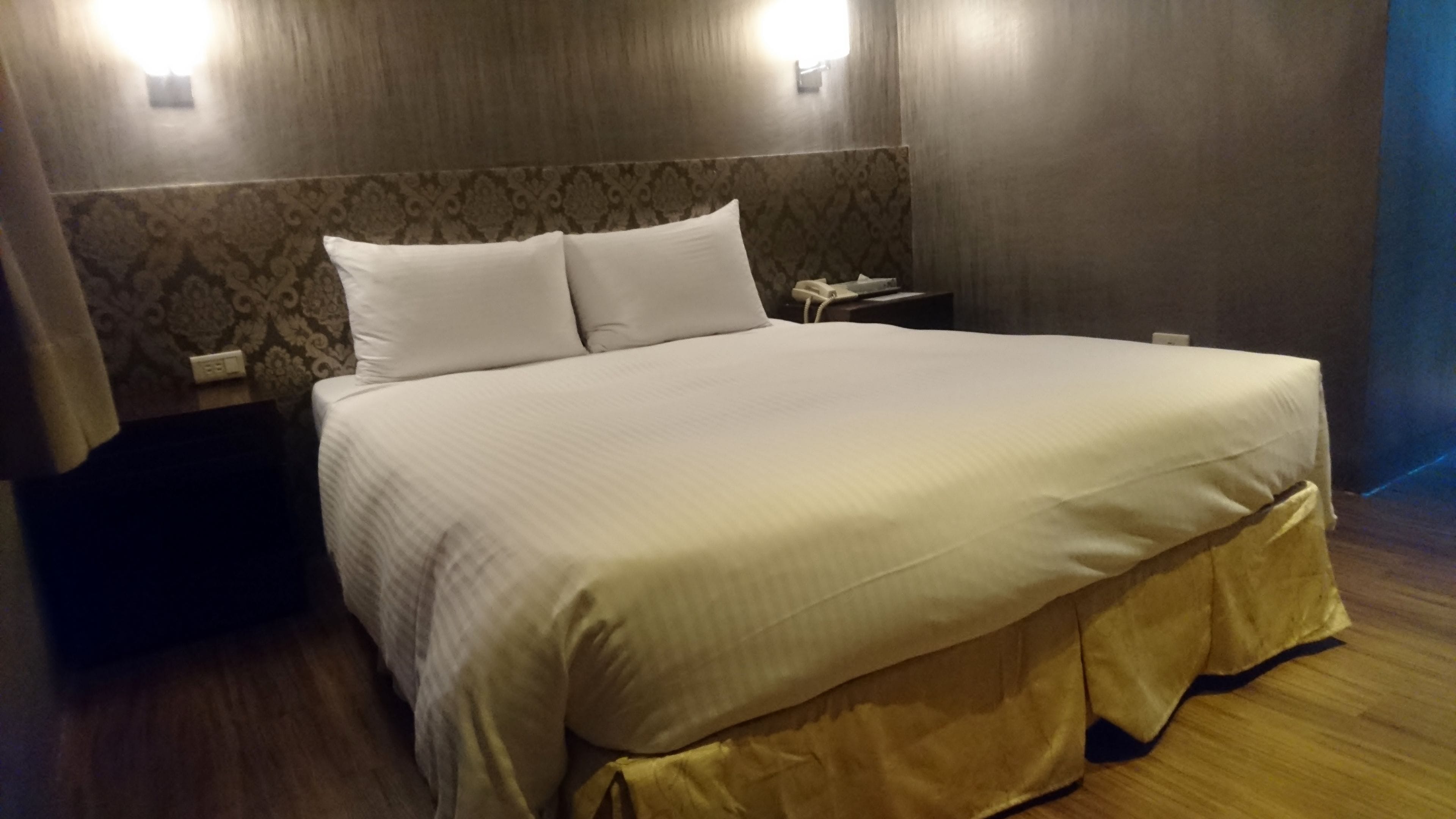 Een netjes opgemaakt bed met een witte deken en twee kussens, een nachtkastje met een lamp, en een behang met een patroon.