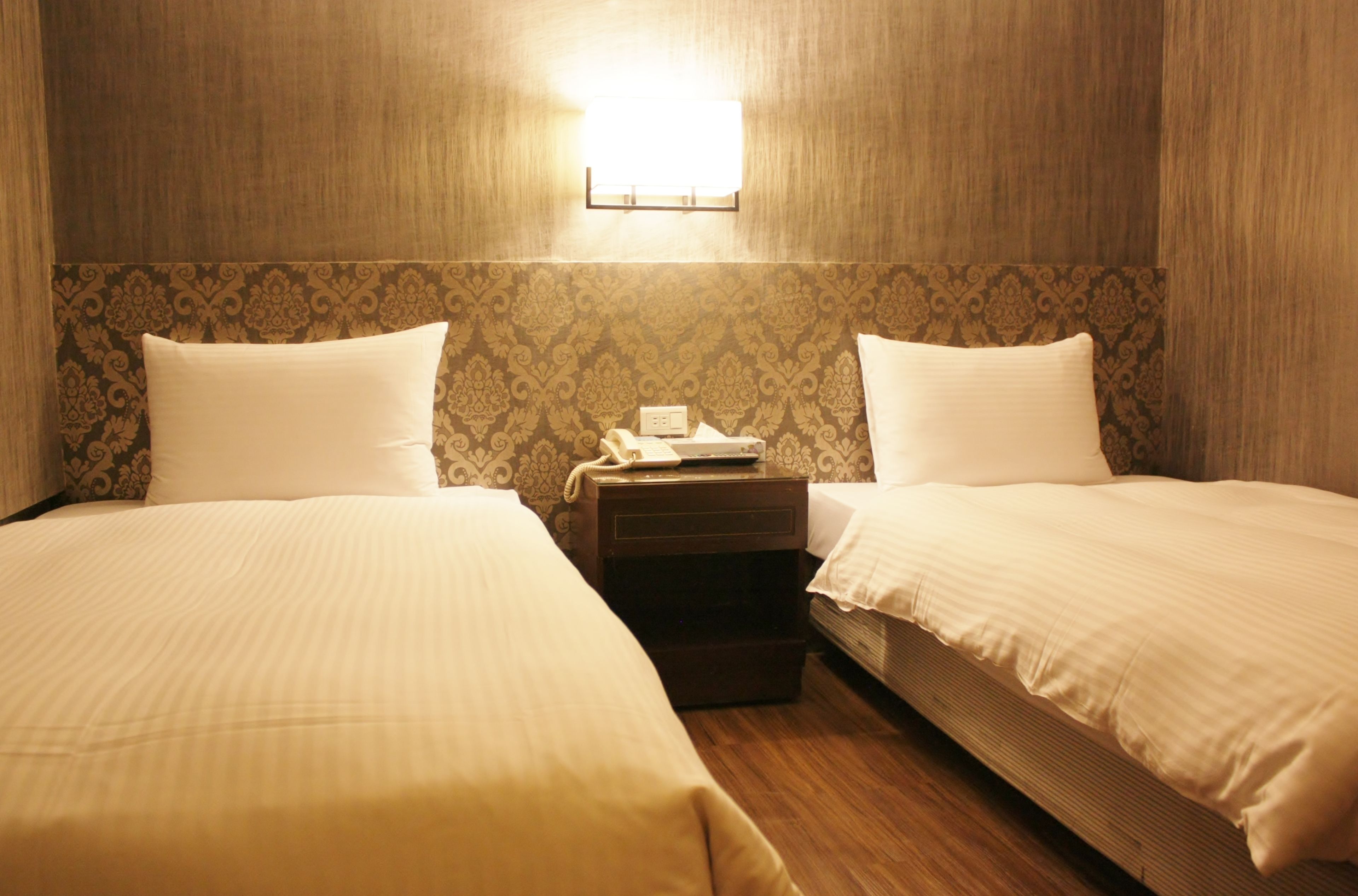 A hotel room with two single beds, a nightstand with a telephone, and a wall-mounted light fixture.