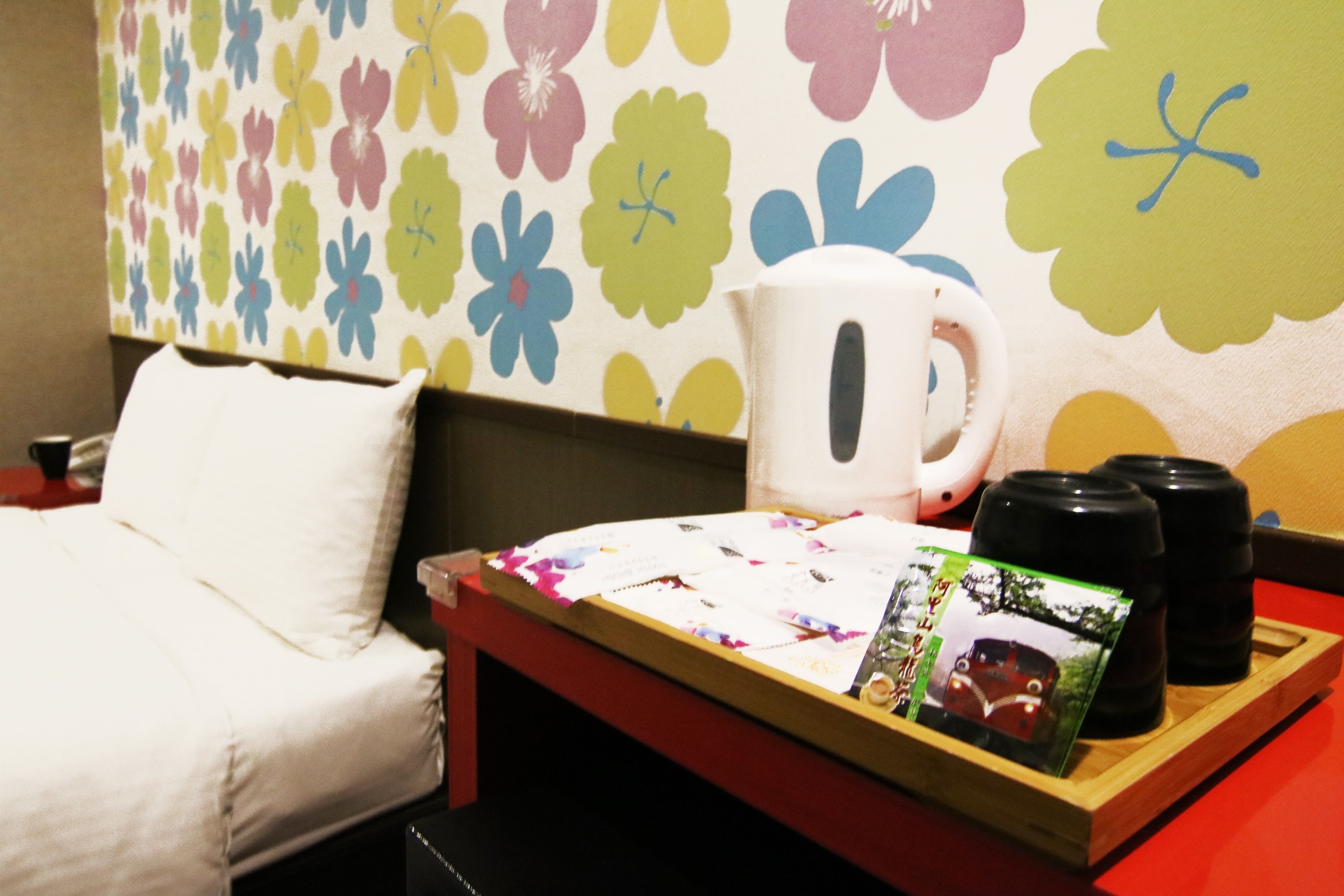 A hotel room with a bed, a bedside table with a kettle, cups, and a tray with a magazine and a photo.