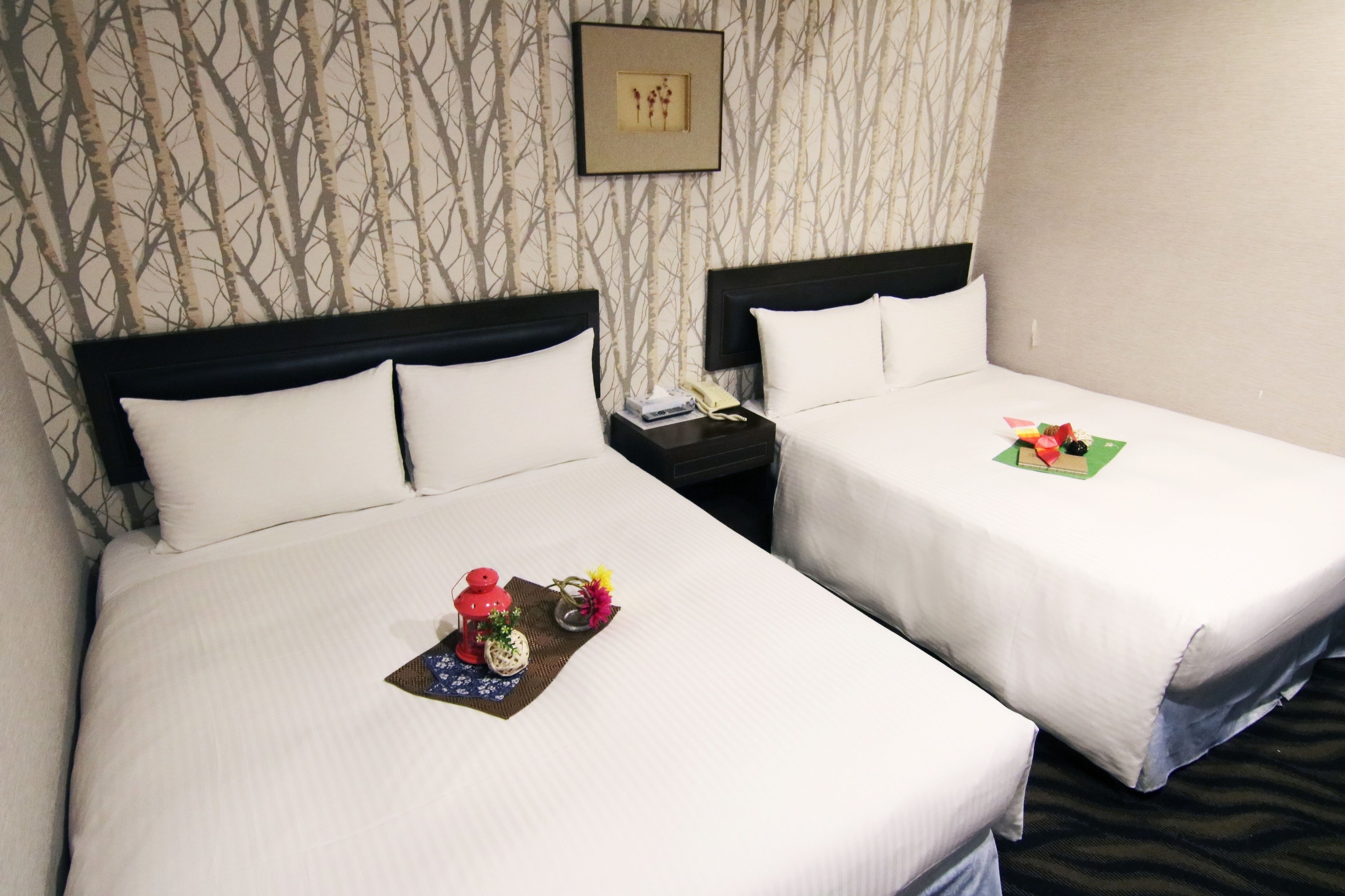 A hotel room with two beds, each with white linens and a headboard. There is a framed picture on the wall above the beds, a nightstand with a telephone, and a small table with a red lantern and flowers.