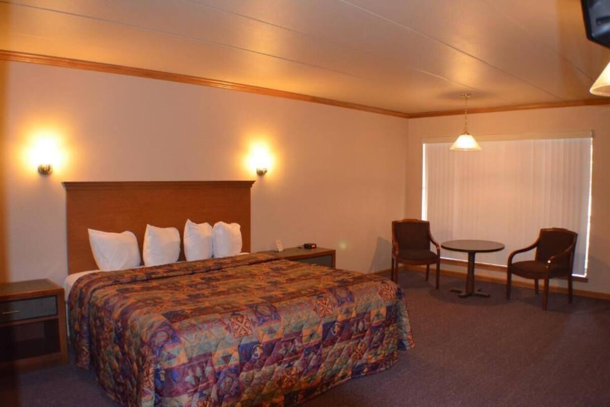 A hotel room with a large bed, two bedside tables, a small round table with two chairs, and a window with blinds.