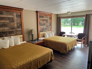 A hotel room with two beds, a table, chairs, a lamp, and a view of the outdoors.