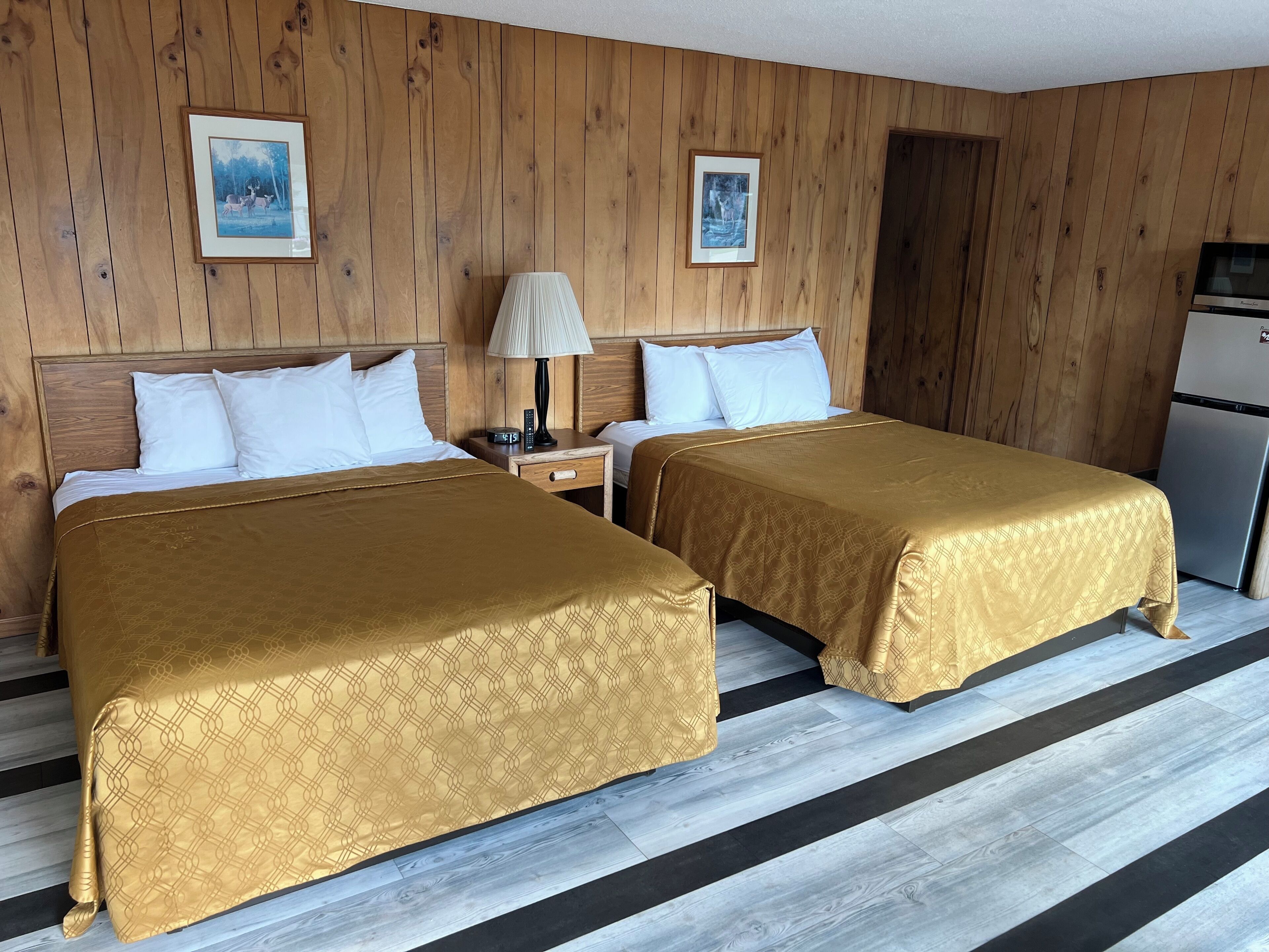 A room with two beds, wooden walls, a small table with a lamp, and a refrigerator.