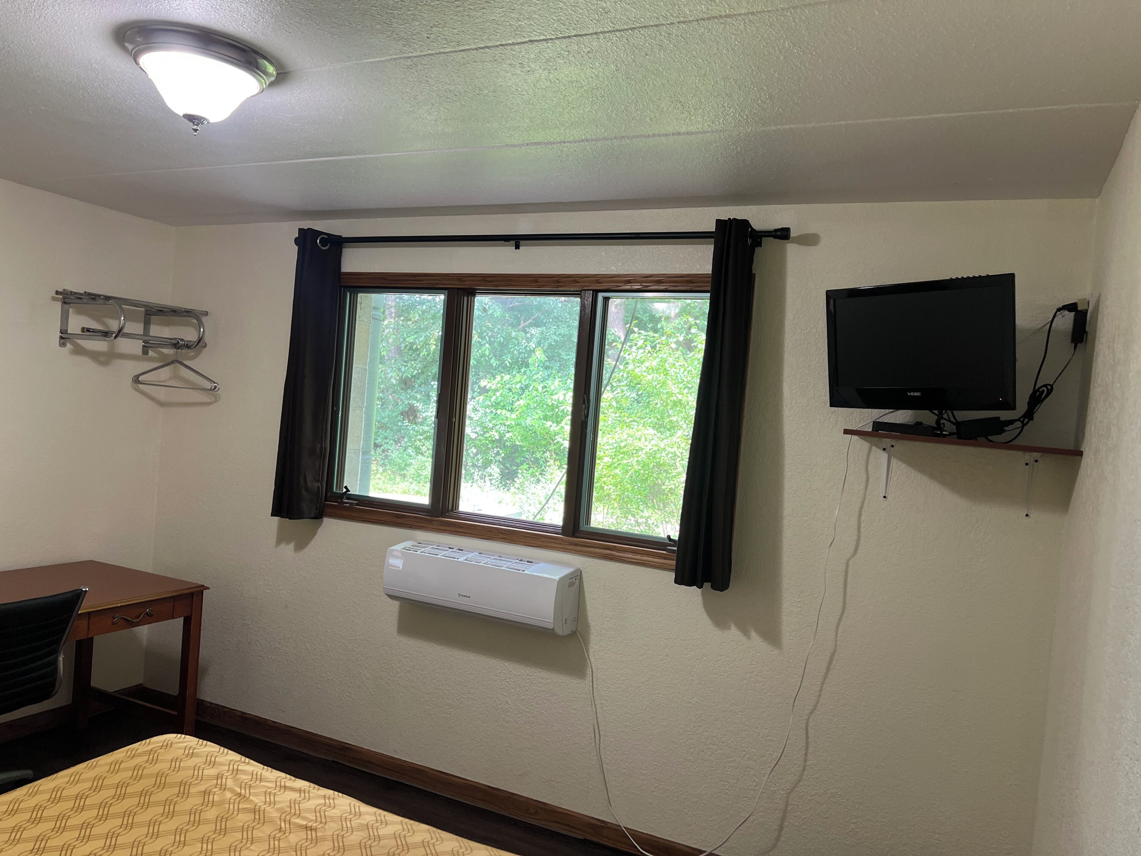 Une chambre comprenant un lit, un bureau, une fenêtre avec des rideaux, un téléviseur fixé au mur et un climatiseur fixé au mur.