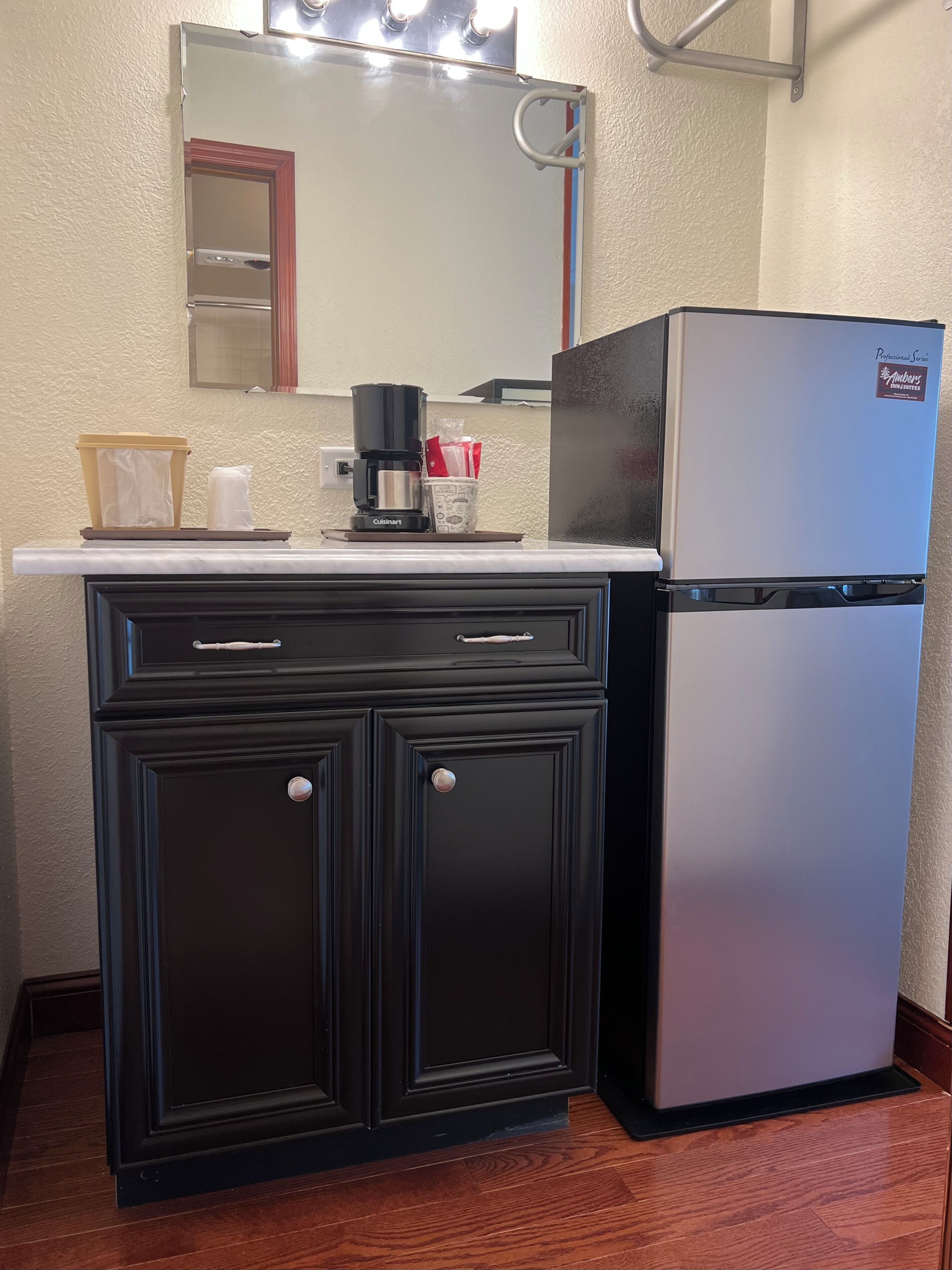 A kitchenette with a stainless steel refrigerator, a coffee maker, a trash can, and a mirror above the counter.