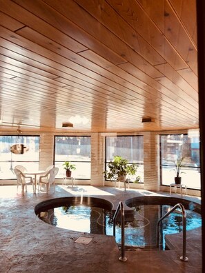 A modern indoor pool area with wooden ceiling beams, large windows, and a seating area with chairs and potted plants. - Amber's Inn & Suites (Wisconsin Dells)