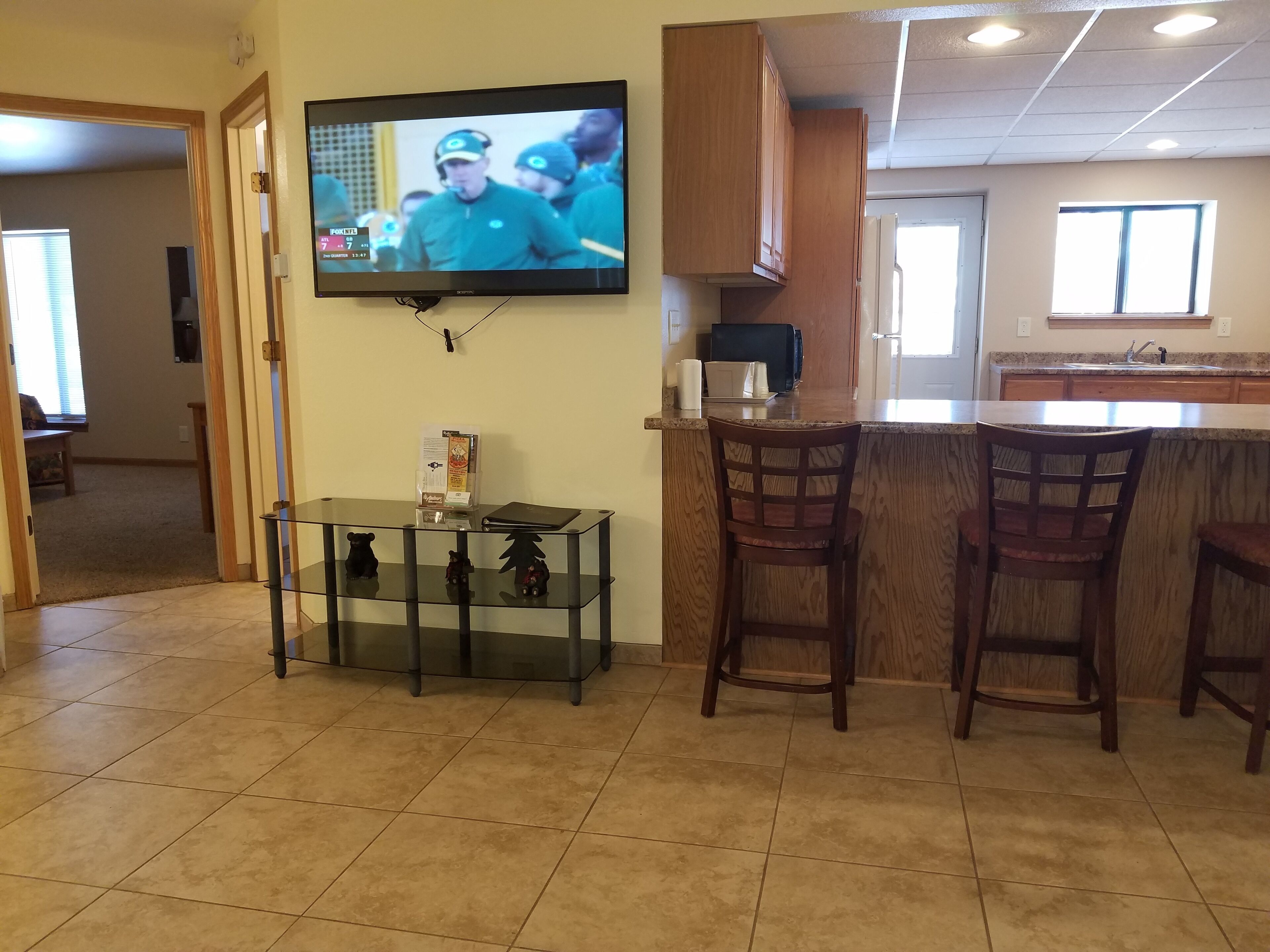 A modern kitchen with a TV mounted on the wall, a glass-top dining table, wooden bar stools, and a kitchen island with a coffee machine.