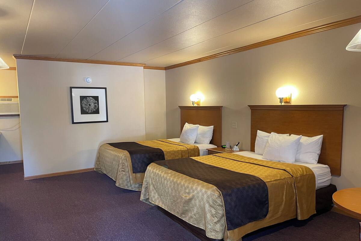 A hotel room with two beds, a small table, a framed picture on the wall, and a ceiling light.
