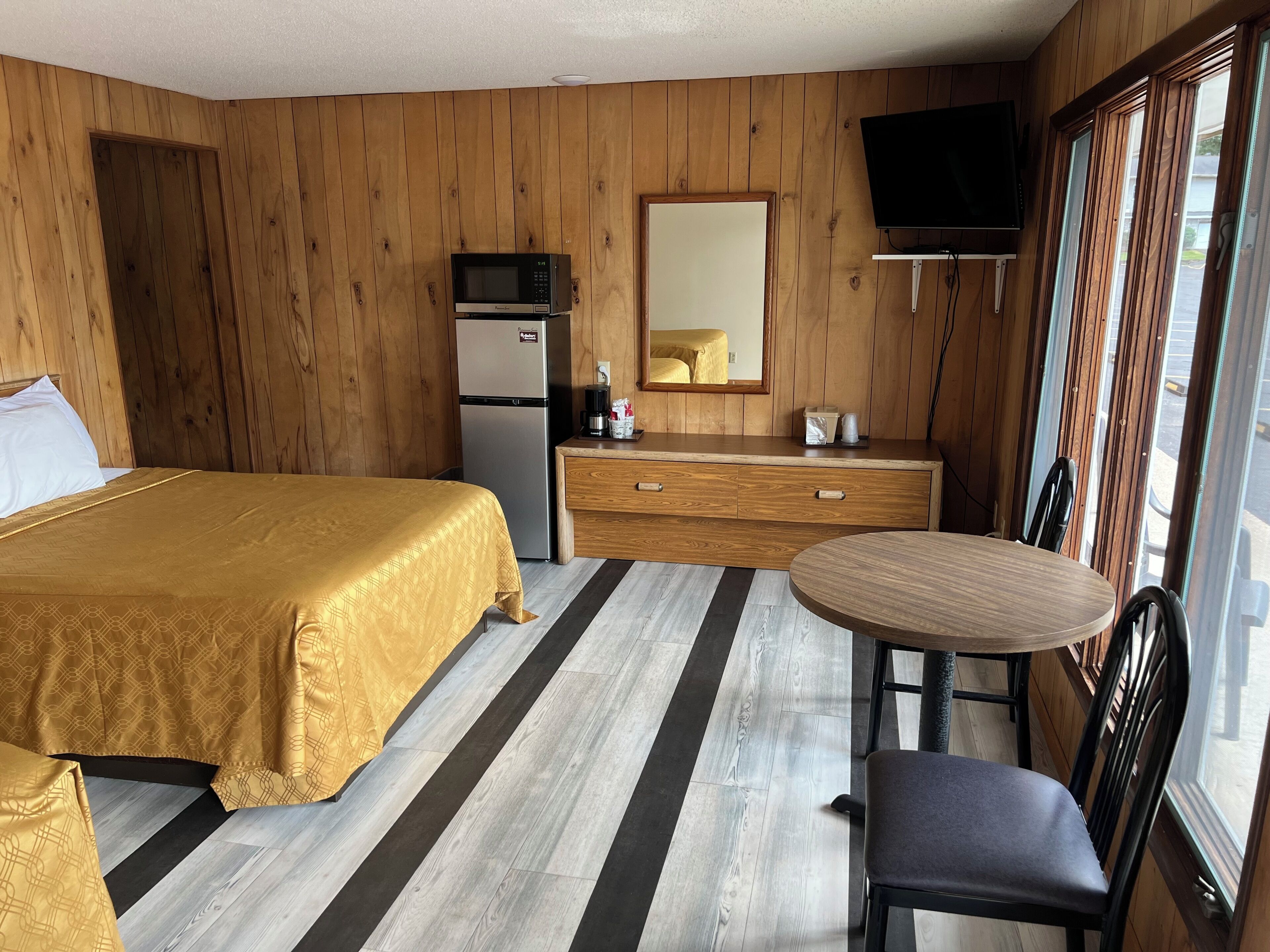 A hotel room with a bed, a small kitchenette, a dining table with a chair, and a TV mounted on the wall.