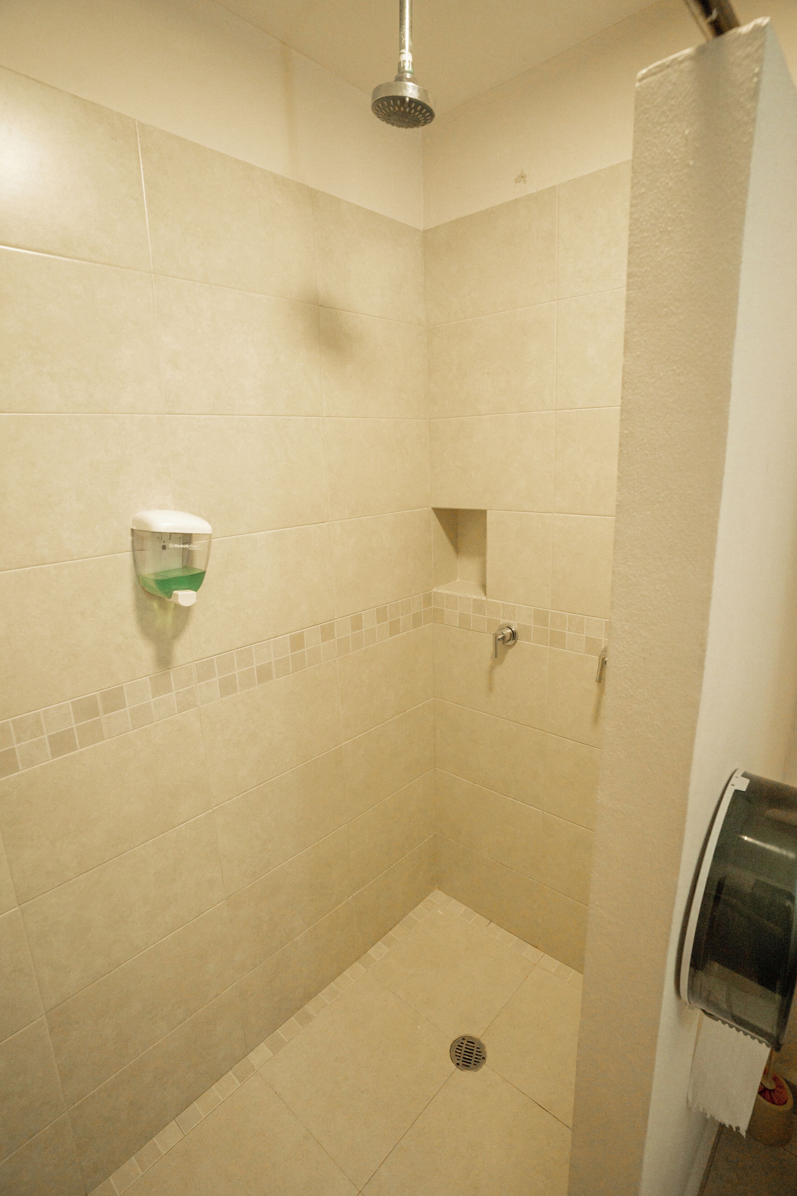 Un espacio de ducha pequeño y cerrado con azulejos color beige, un cabezal de ducha manual y un dispensador de jabón en la pared.