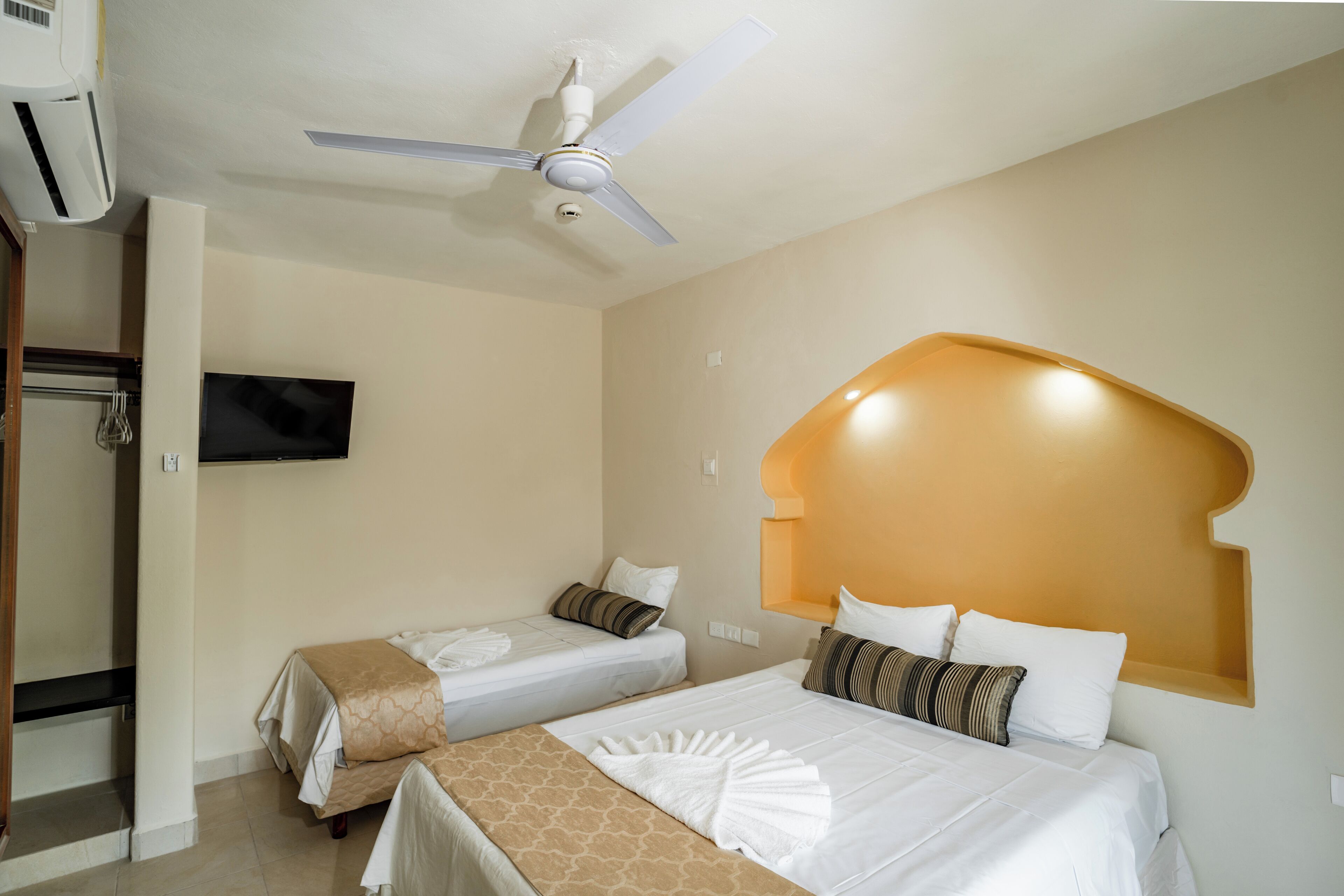 Une chambre d’hôtel avec deux lits, un ventilateur de plafond, un téléviseur fixé au mur et une tête de lit en bois.