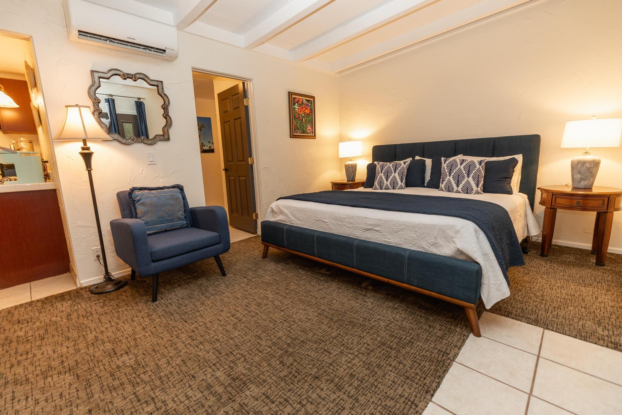 A bedroom with a large bed, two bedside tables with lamps, a chair, a mirror, and a door leading to another room.