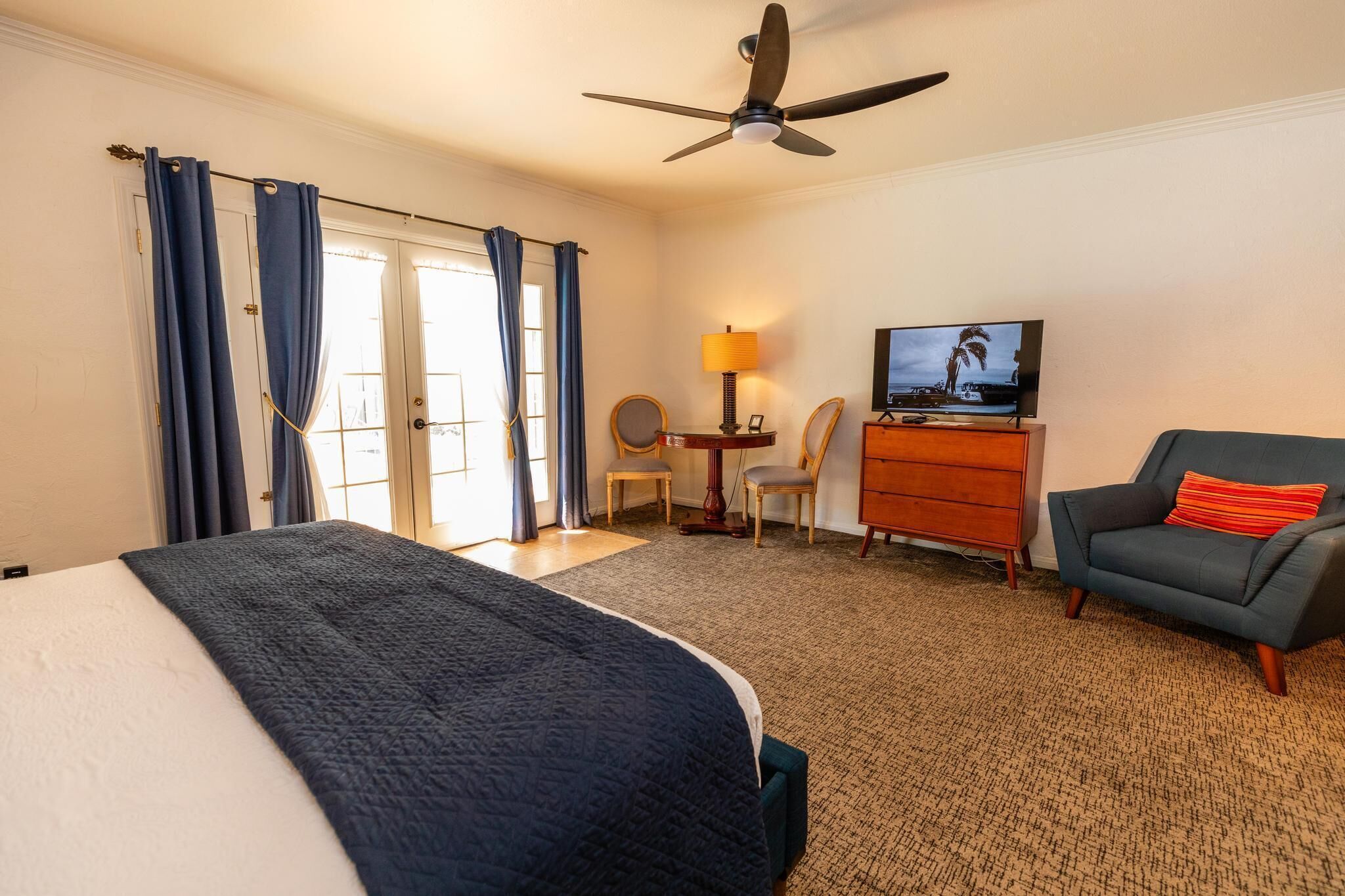 Een hotelkamer met een bed, een bureau met lamp, een televisie, een stoel en een plafondventilator.