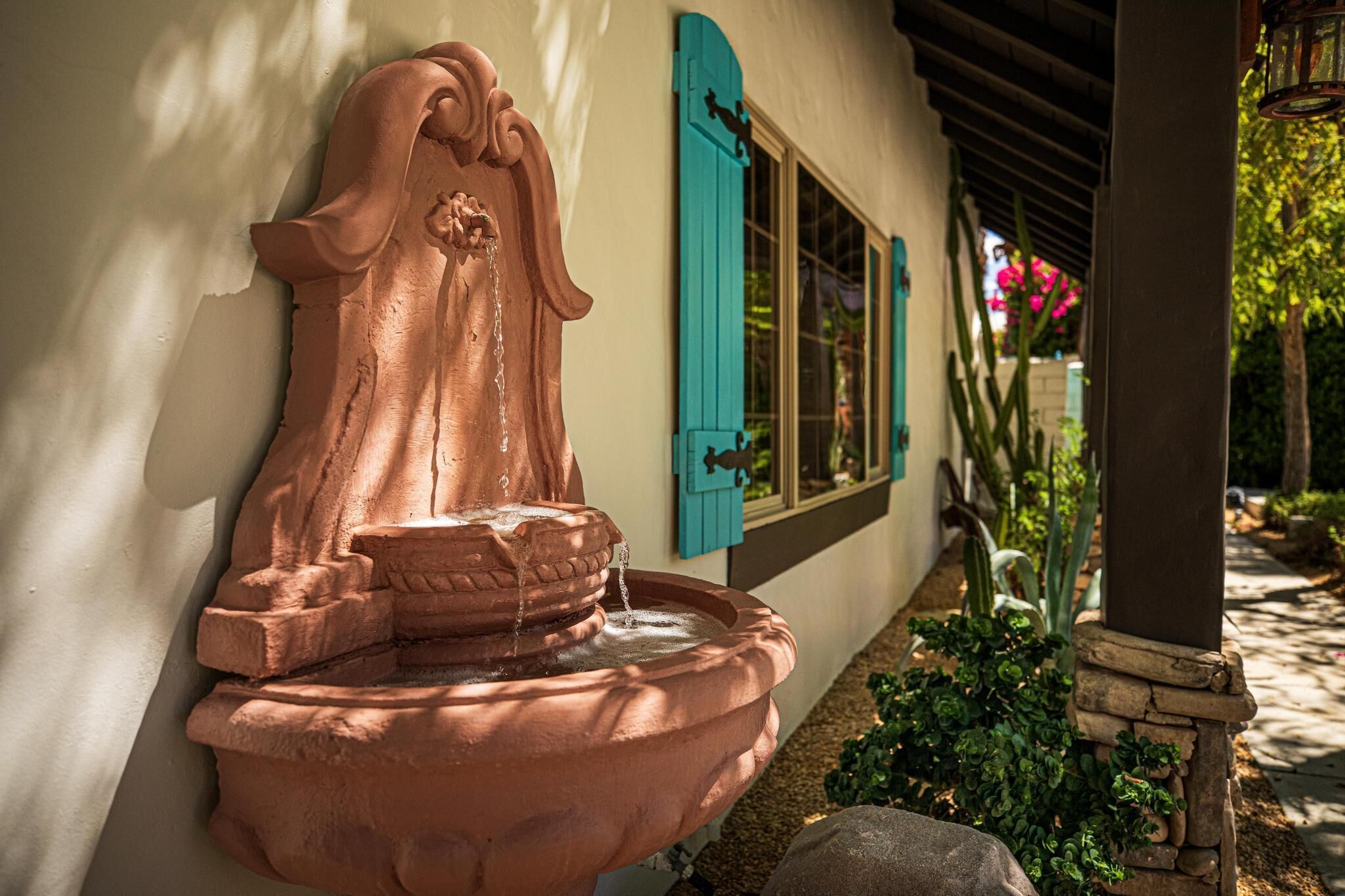 A terracotta fountain with water flowing, attached to a building with turquoise shutters and a stone pillar.