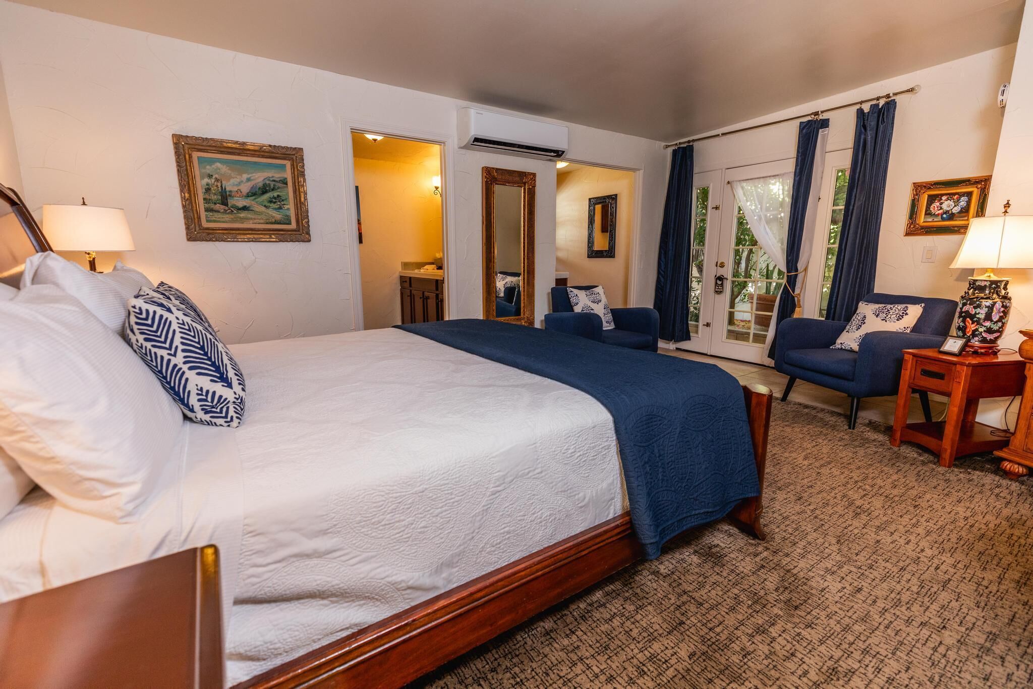 A hotel room with a bed, a chair, a small table, and a mirror. There is a door leading to a bathroom and a window with curtains.