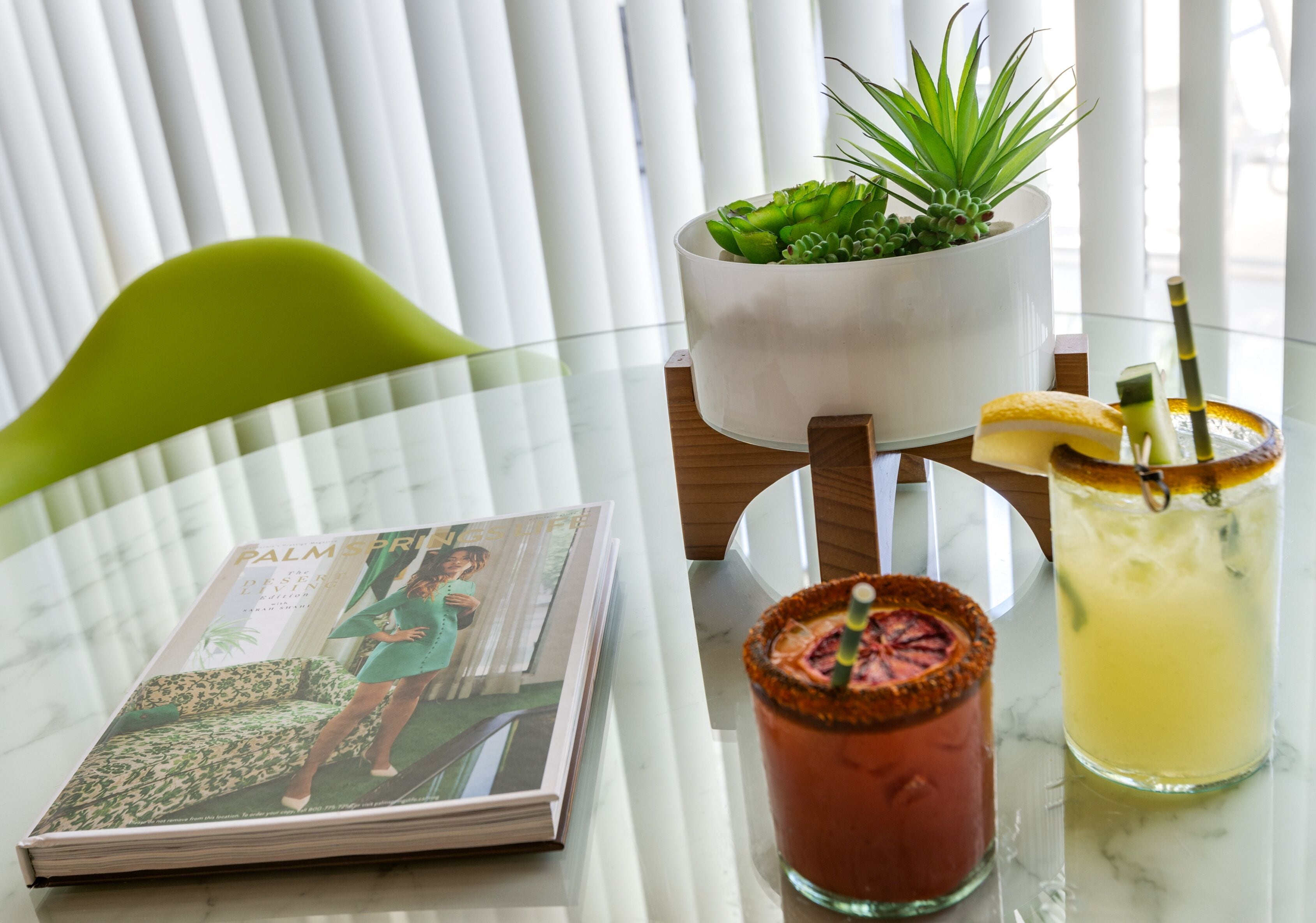 Un magazine sur une table, une boisson décorée d’une garniture et une plante dans un pot blanc.