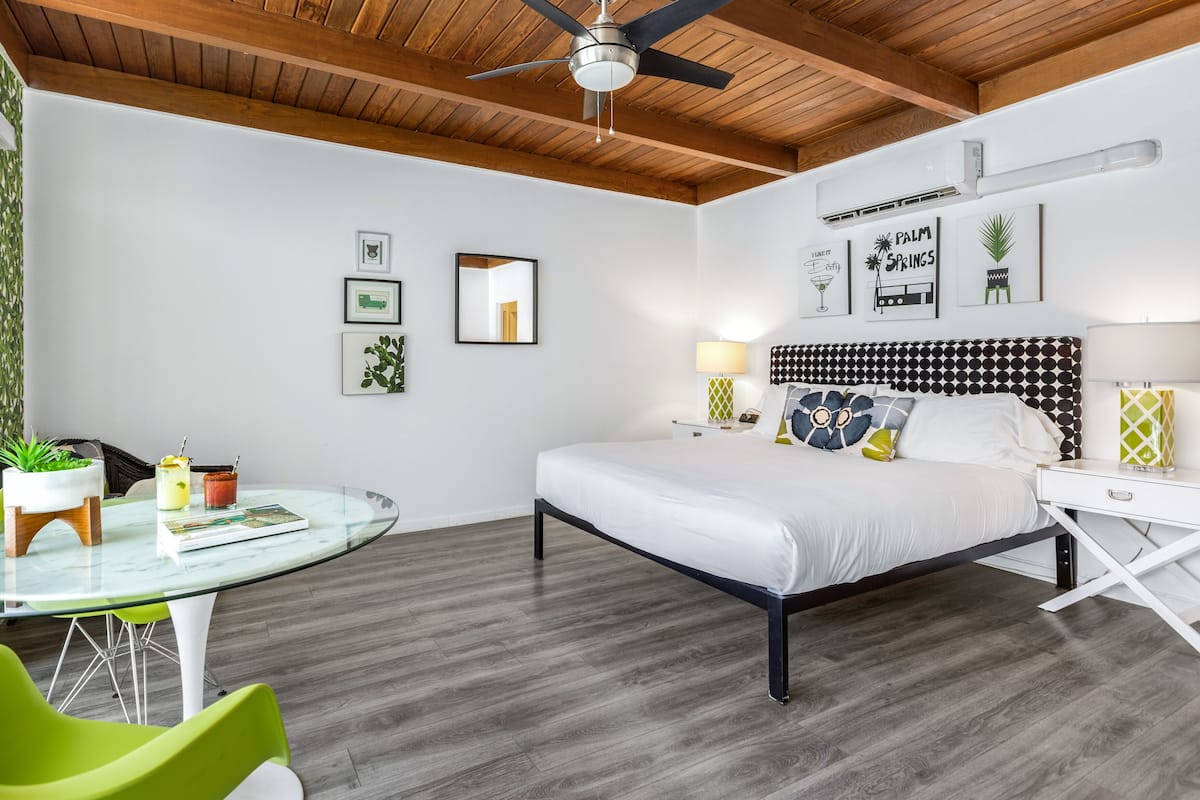 A modern hotel room with a bed, a glass table, green chairs, a ceiling fan, and wall art.