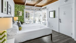 A bedroom with a bed, a desk, and a chair. There is a window with blinds, a ceiling fan, and a door with a lock. - The Three Fifty Hotel (Palm Springs)
