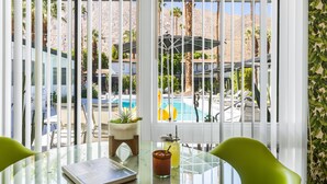 A room with a glass table, a book, a drink, and a view of a pool and palm trees. - The Three Fifty Hotel (Palm Springs)