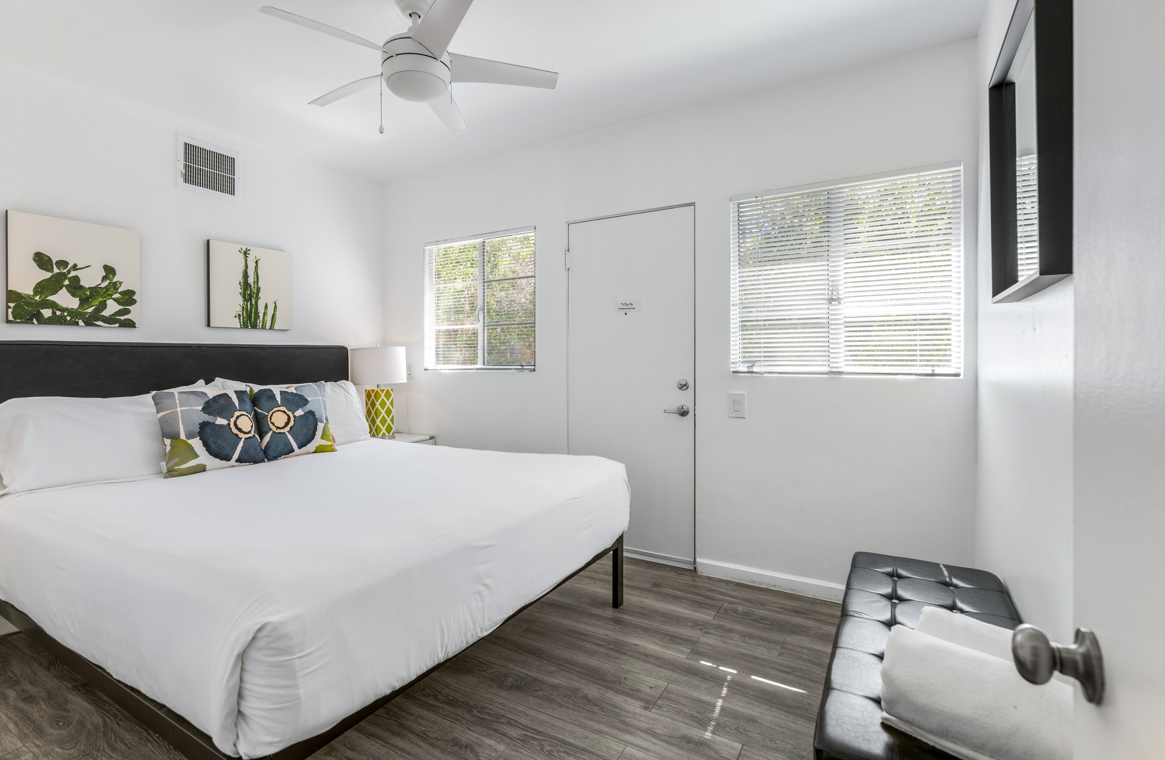 Un dormitorio con cama, ventilador de techo, dos ventanas con persianas, una puerta y un pequeño banco.