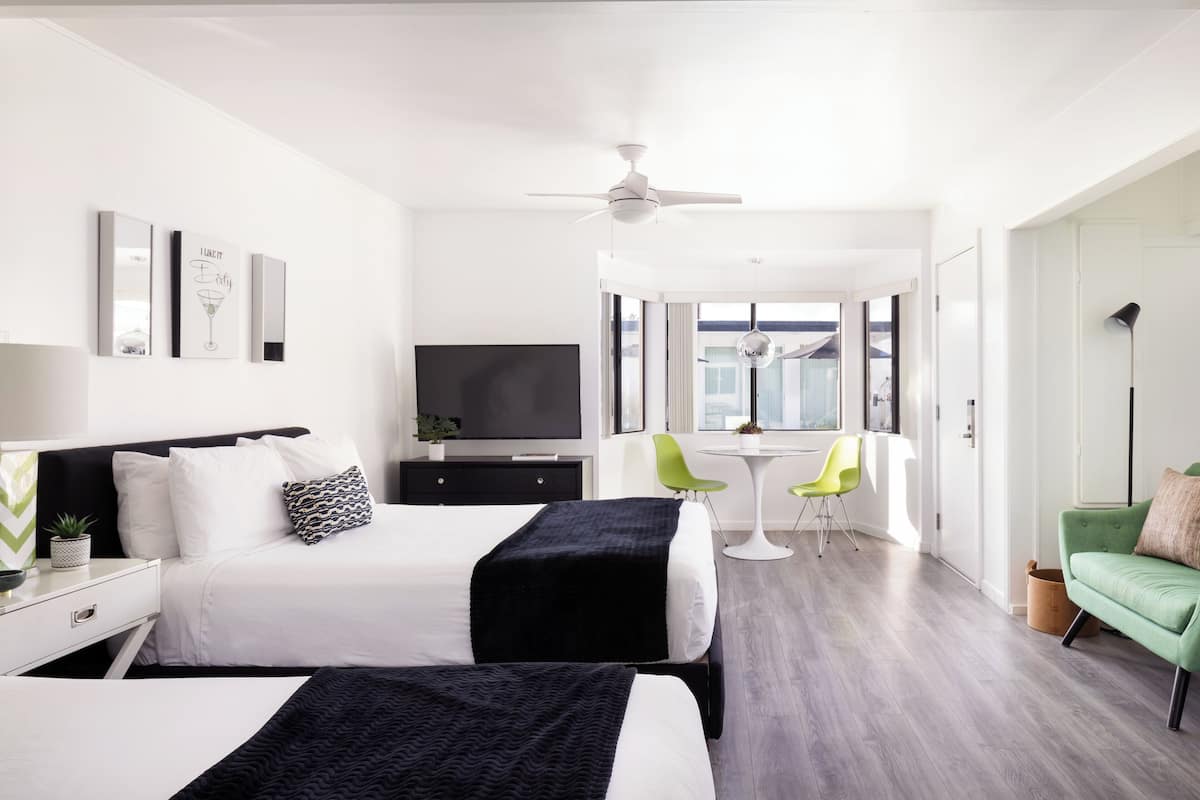 A modern hotel room with two beds, a flat-screen TV, a ceiling fan, a dining area with a table and chairs, and a green sofa.