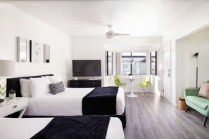 A modern hotel room with two beds, a flat-screen TV, a ceiling fan, a dining area with a table and chairs, and a green sofa.